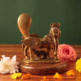 Brass Kamadhenu idol back view with calf beneath, on a wooden platform with white and pink flowers, green backdrop.
