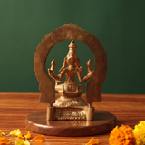 Lakshmi idol in back view with circular arch prabhavali seated on a lotus base with yellow and orange marigolds on a wooden plate.