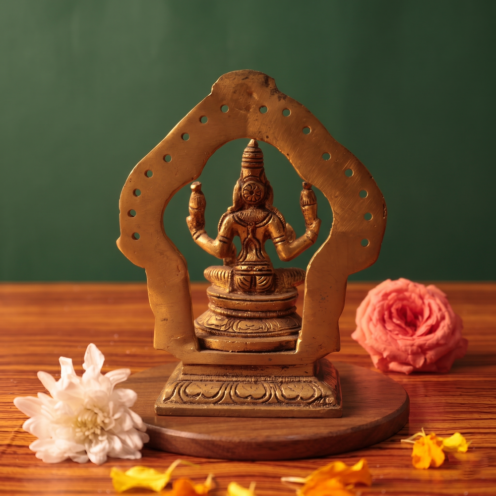 Brass Lakshmi idol back view, seated within a simple perforated arch, on a wooden platform with white and pink flowers, green backdrop.
