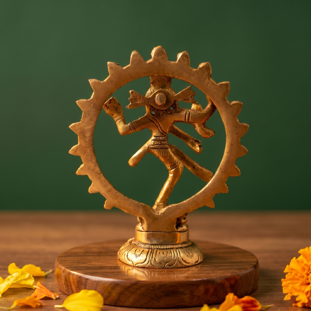 Brass Nataraja idol back view dancing within a fire ring, on a wooden platform with marigold flowers, green backdrop.
