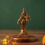 Brass Meenakshi idol back view, standing with a parrot, on a wooden platform with scattered marigold petals, green backdrop.
