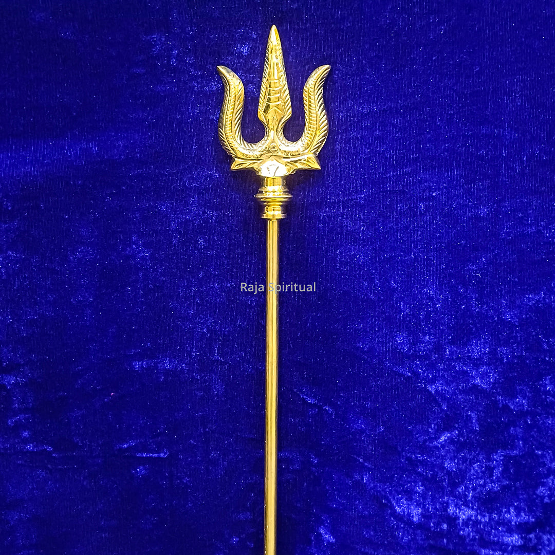 Brass muppattai sulam trishul 3ft with large feather-edged trident head shown in straight upright front view against blue velvet backdrop.