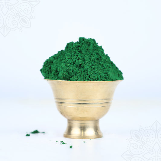 Green kumkum powder heaped in a brass bowl front-facing view with small powder spill beside it against a light blue background