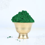 Green kumkum powder heaped in a brass bowl front-facing view with small powder spill beside it against a light blue background