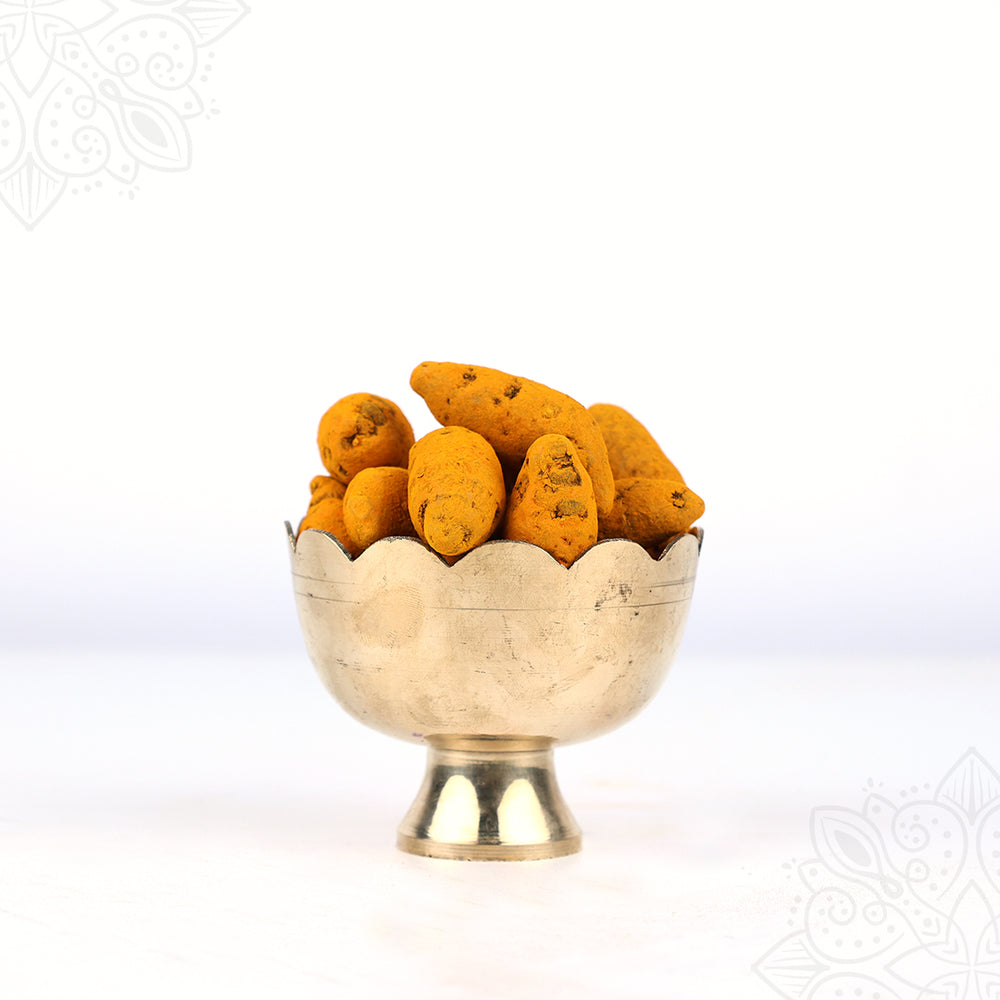 Raw manjal kizhangu turmeric roots filled in a scallop-edged puja bowl shown in straight front view with full base visible against white backdrop.
