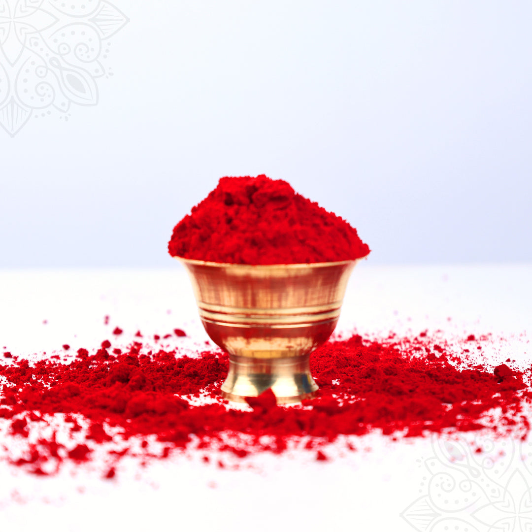Red kumkum powder heaped in a brass puja bowl, spilled on white surface, front view with mandala backdrop.