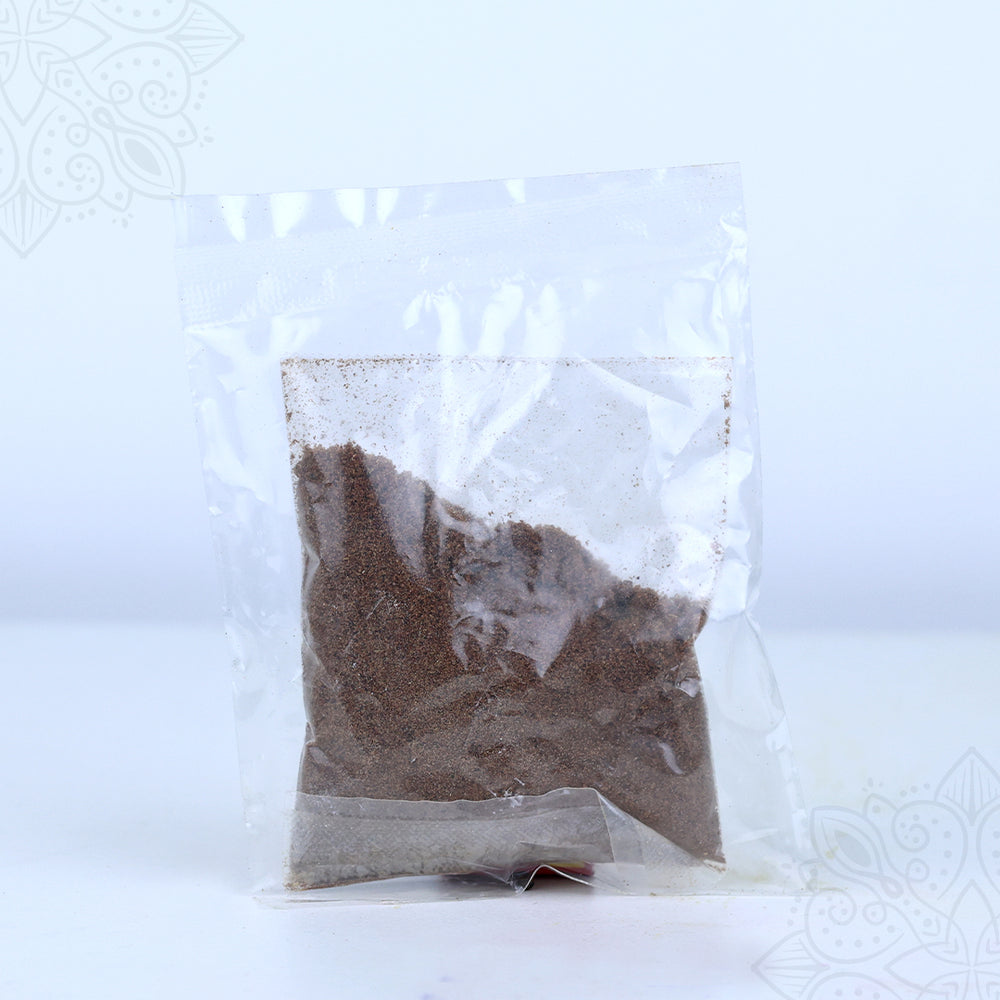 Thirtha parimalam powder in a clear sealed plastic packet showing dark brown aromatic powder in straight front view against white backdrop.